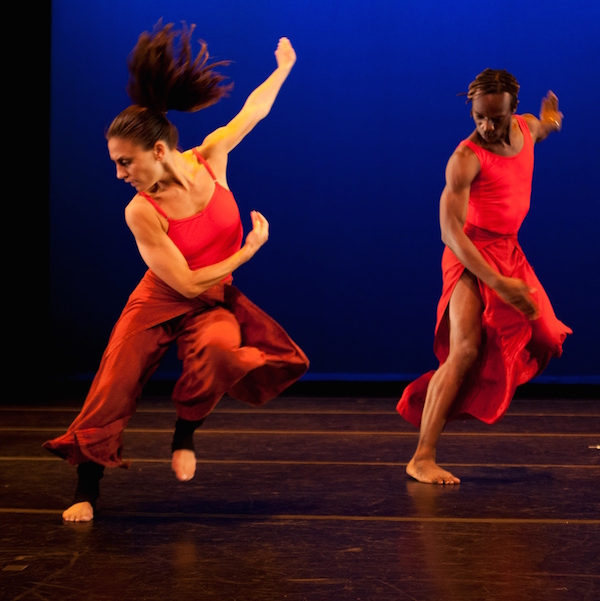 Two dancers in red costumes in a flurry of dynamic movement. One dancer's pony tail is lifted into the air.